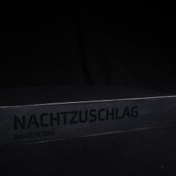 ein schwarz schimmernder Schuber welcher den Titel Nachtzuschlag ebenfalls in schwarz ziert. In diesem Schuber befinden sich elf ebefalls schwarze Magazine.