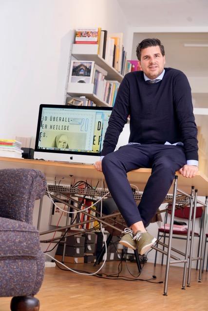 Jens Müller sitzt auf dem Tisch. Neben ihm ist ein Mac. Hinter dem Tisch steht ein Buchregal mit vollen Büchern.