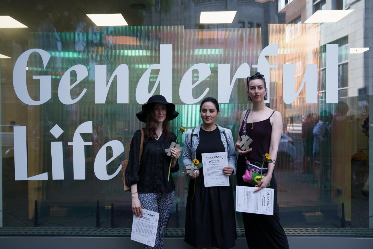Drei Personen stehen vor einem halbtransparenten Schaufenster mit dem Schriftzug &amp;quot;Genderful Life&amp;quot;. In den Händen halten sie je eine Urkunde, eine Sonnenblume und ein graues Ungleichzeichen.