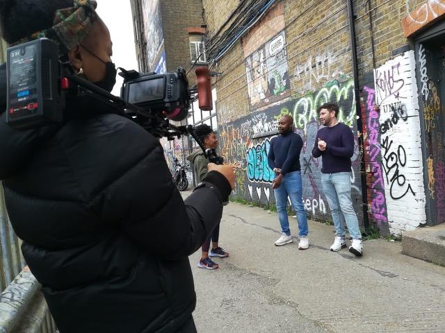 Eine Filmemacherin filmt zwei Männer, die vor einer mit Graffiti bedeckten Backsteinwand in einer städtischen Gasse stehen. Die Filmemacherin trägt eine schwarze Daunenjacke und ein Kopftuch und hält eine professionelle Kameraausrüstung. Die beiden Männer, einer in einem dunkelblauen Pullover und Jeans, der andere in einem dunklen Pullover und hellen Jeans, unterhalten sich. Eine dritte Person, eine Frau in einer schwarzen Jacke, steht in der Nähe.