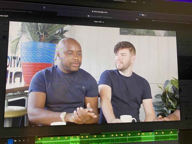 Ein Videoschnittprogramm zeigt zwei Männer, die zusammen in einer entspannten Umgebung sitzen. Beide haben Kaffeetassen vor sich. Der Mann im dunkelblauen T-Shirt spricht gestikulierend, während der andere im schwarzen T-Shirt ihm aufmerksam zuhört. Im Hintergrund befinden sich eine bunte Dekoration mit Streifen und grüne Pflanzen. Die Benutzeroberfläche des Schnittprogramms mit Videotimeline und Audiowellenform ist sichtbar.