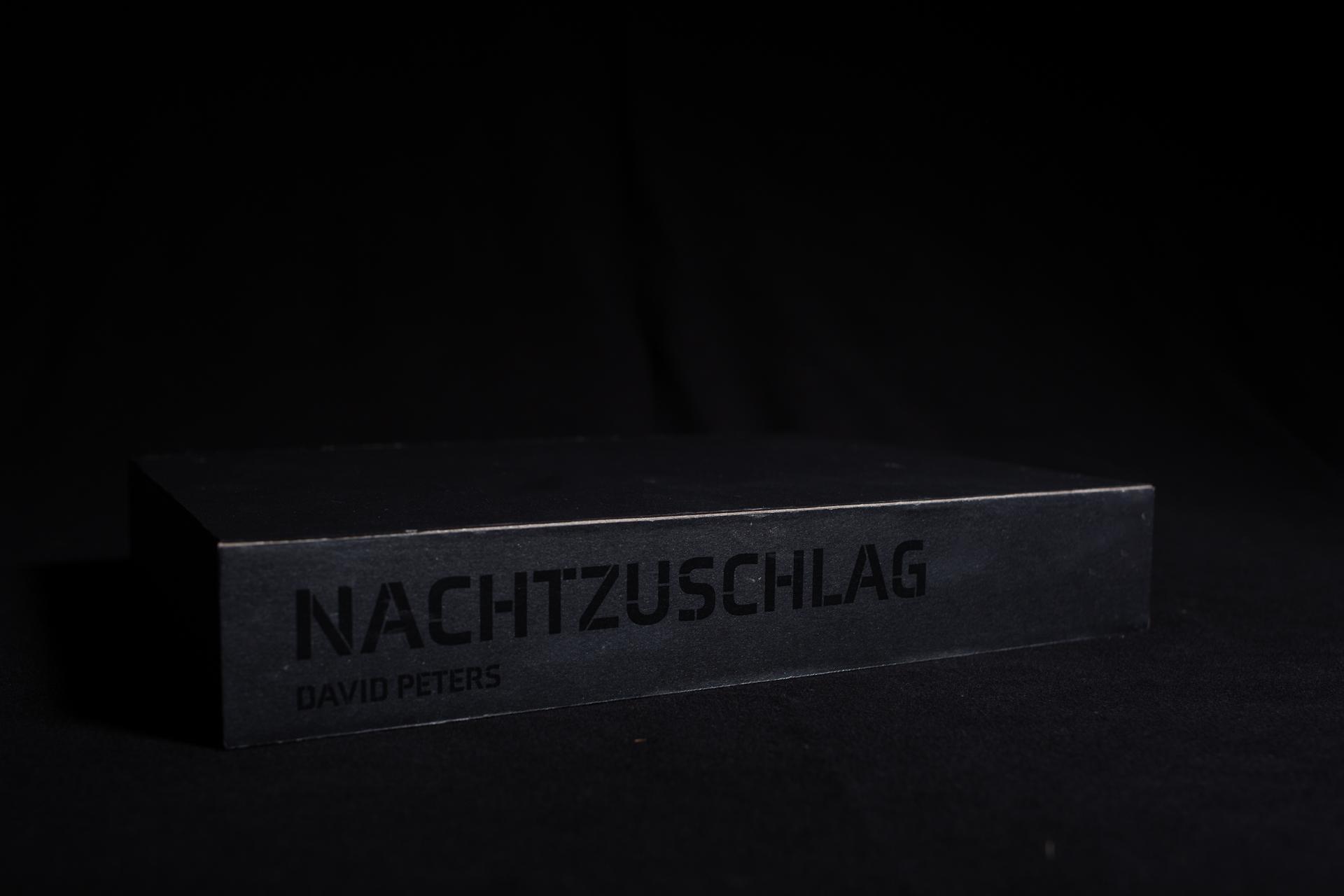 ein schwarz schimmernder Schuber welcher den Titel Nachtzuschlag ebenfalls in schwarz ziert. In diesem Schuber befinden sich elf ebefalls schwarze Magazine.